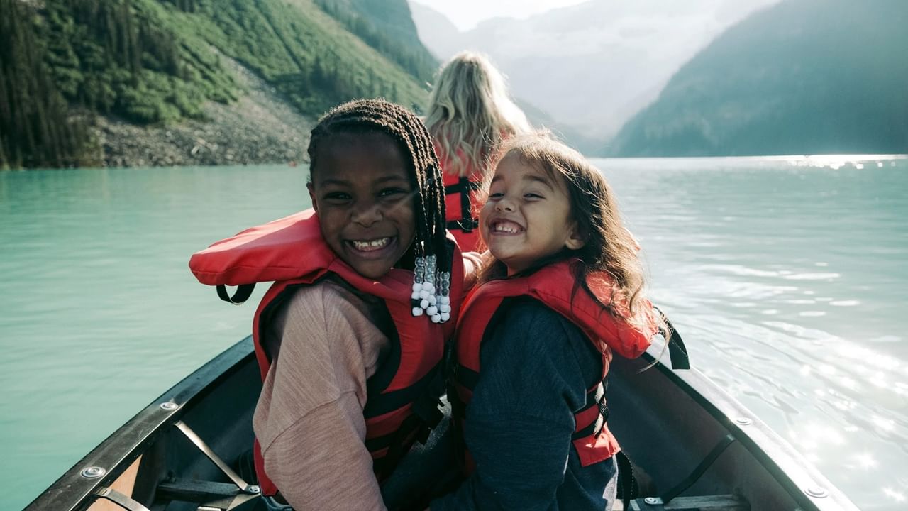 Two girls in life jackets smiling and sitting in the back of a canoe on a lake.