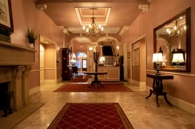 Elegant hallway with chandelier, fireplace, red rugs, and a large mirror reflecting chandelier and lamp.