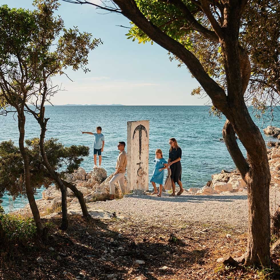 A family of four stands near a white door by the sea, with trees and rocks nearby.