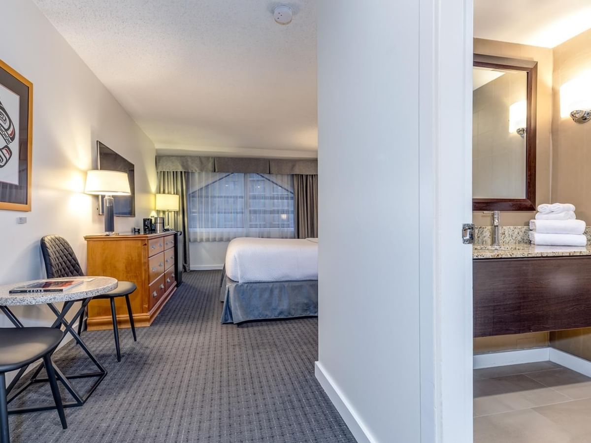 A hotel room with a bed, desk, chairs, dresser, and bathroom with a sink and mirror.