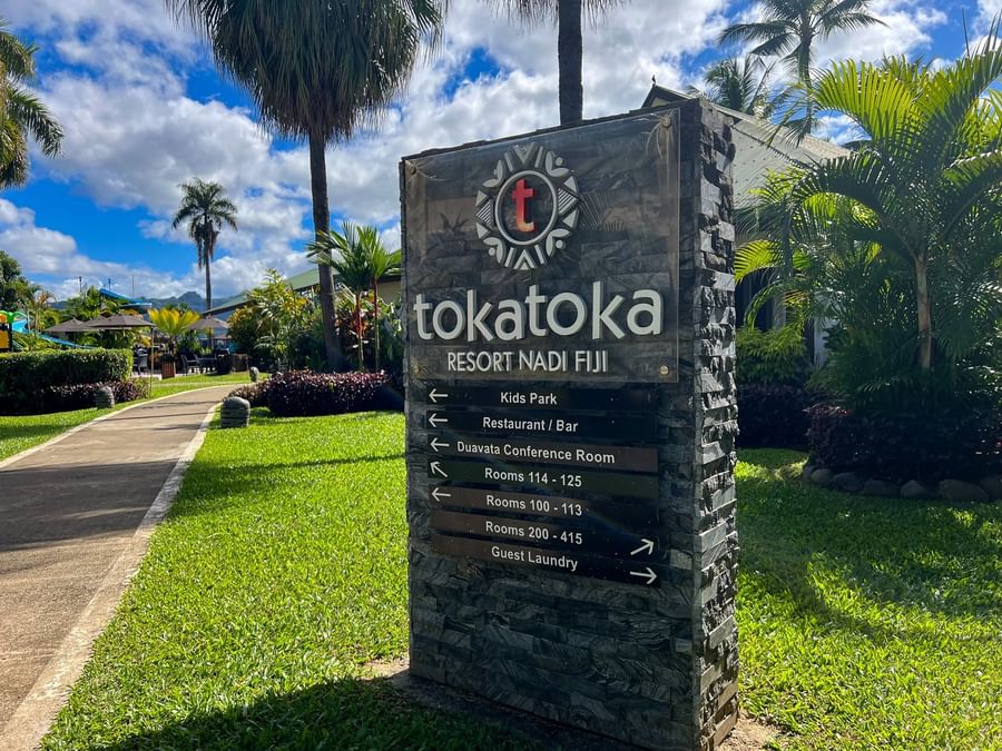 Large stone-textured welcome sign with green roofs by a grassy field surrounding palm trees at TokaToka Resort Nadi Fiji