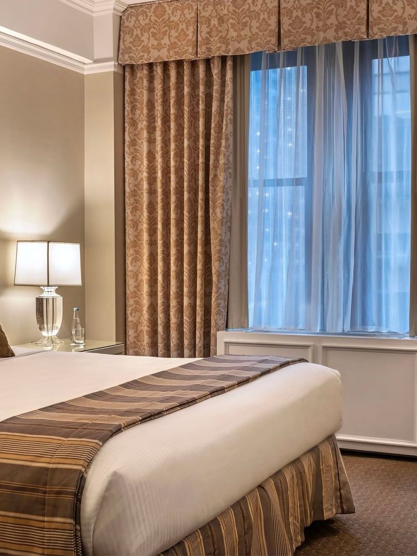 Large bed with white linens and gold-patterned curtains by a bright window in Premier City view suite at Warwick New York