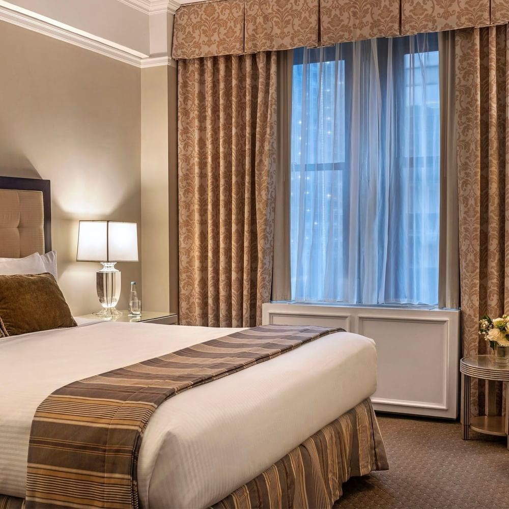 Large bed with white linens and gold-patterned curtains by a bright window in Premier City view suite at Warwick New York