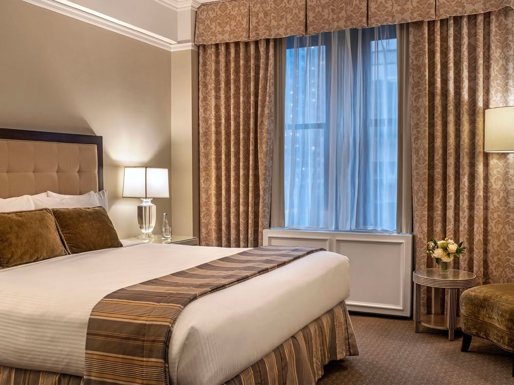 Large bed with white linens and gold-patterned curtains by a bright window in Premier City view suite at Warwick New York