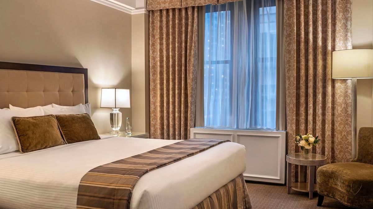 Large bed with white linens and gold-patterned curtains by a bright window in Premier City view suite at Warwick New York