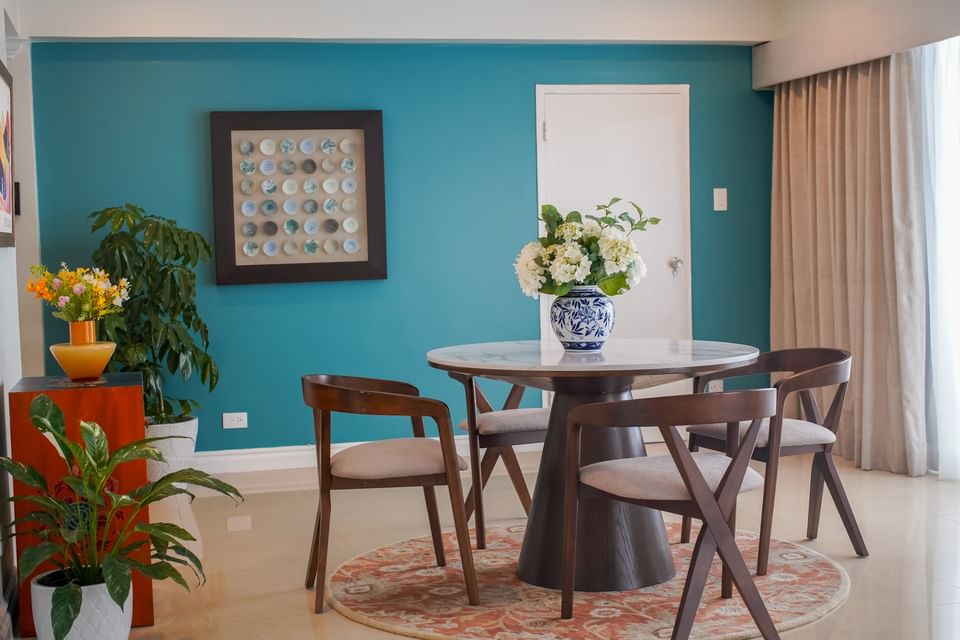 Dining space with flower vase on a round wooden table & four chairs facing a teal wall with art at Jamaica Pegasus Hotel