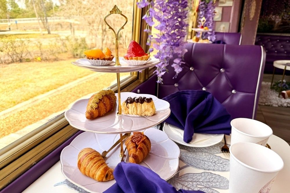 A table on a bus with purple leather seats set with a tiered plate of delicate pastries, blue napkins, and decorated with draped purple flowers on Bloom Ride Orlando.