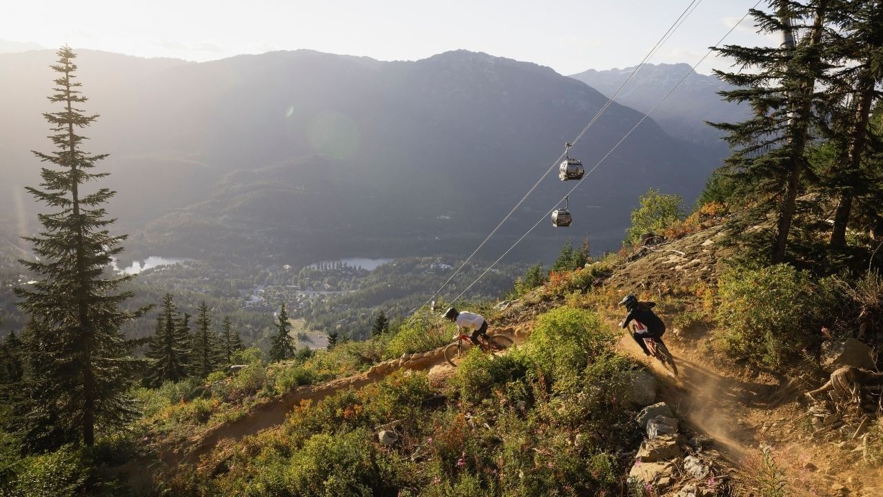 Mountain bikers ride a Whistler trail beneath the Peak 2 Peak Gondola with valley views below.