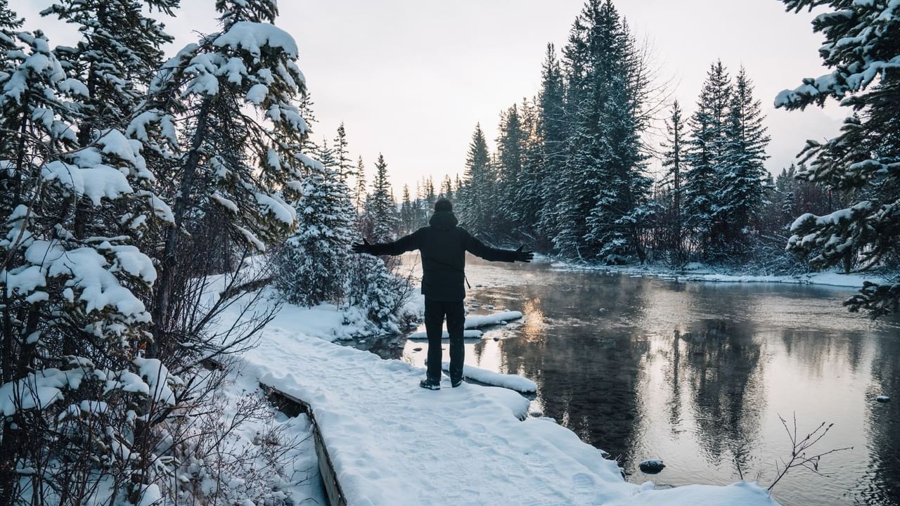 Man standing on a snowy path by a river, surrounded by snow-covered trees.