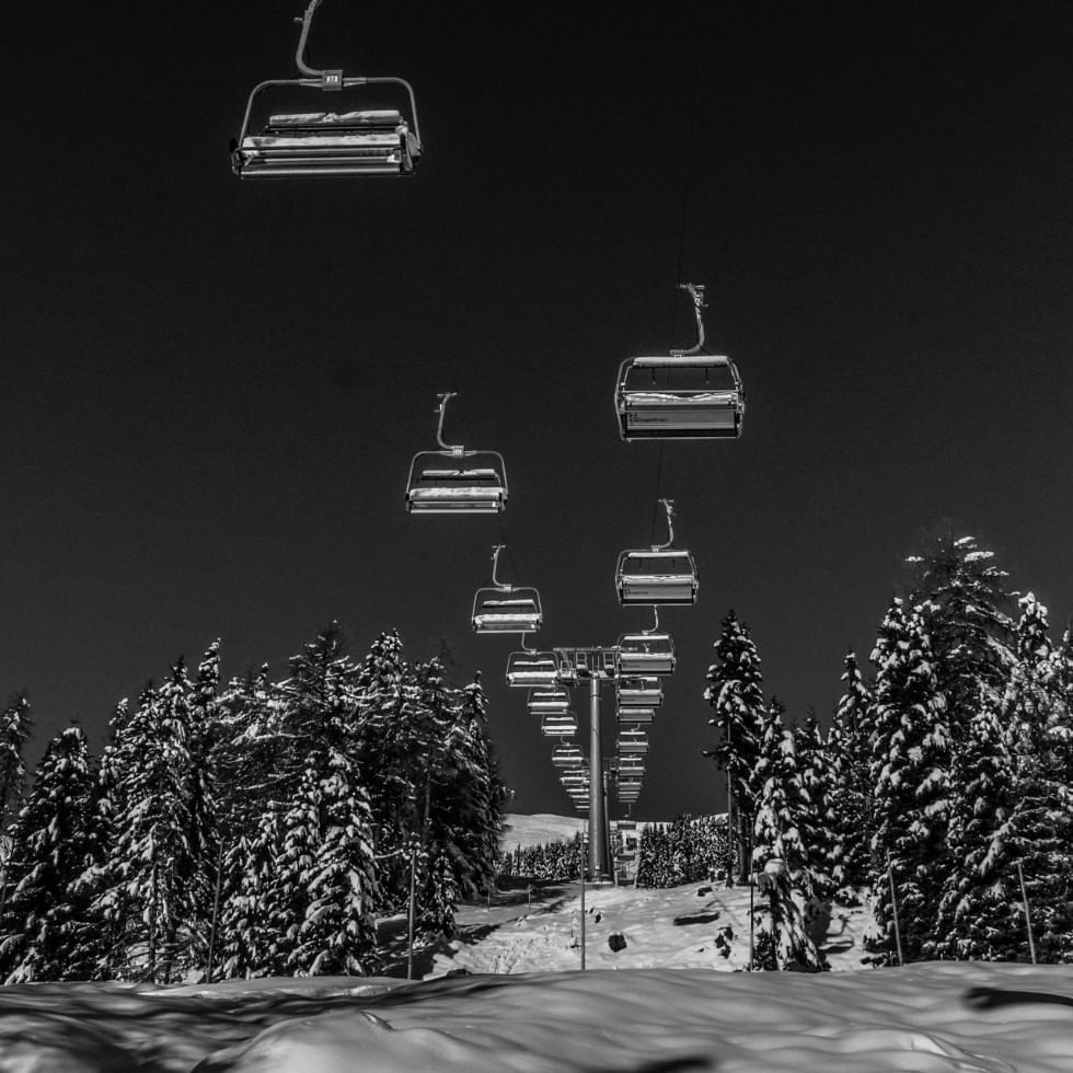 A ski lift with empty chairs over a snowy slope with pine trees, captured in black and white.