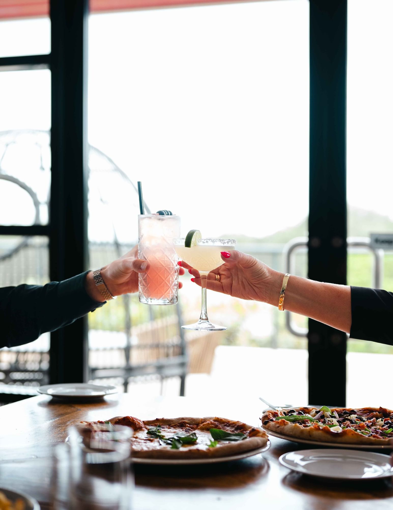 Two people toasting with cocktails near pizzas and plates on a wooden table by a window.