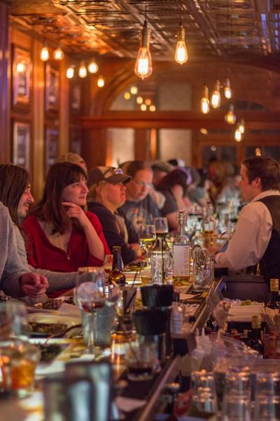 A crowded bar with people sitting at the counter, a bartender serving them, in Cascades Restaurant at The Stanley Hotel