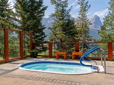 Outdoor hot tub with blue railings sits on a stone patio at Falcon Crest Lodge, surrounded by tall pine trees