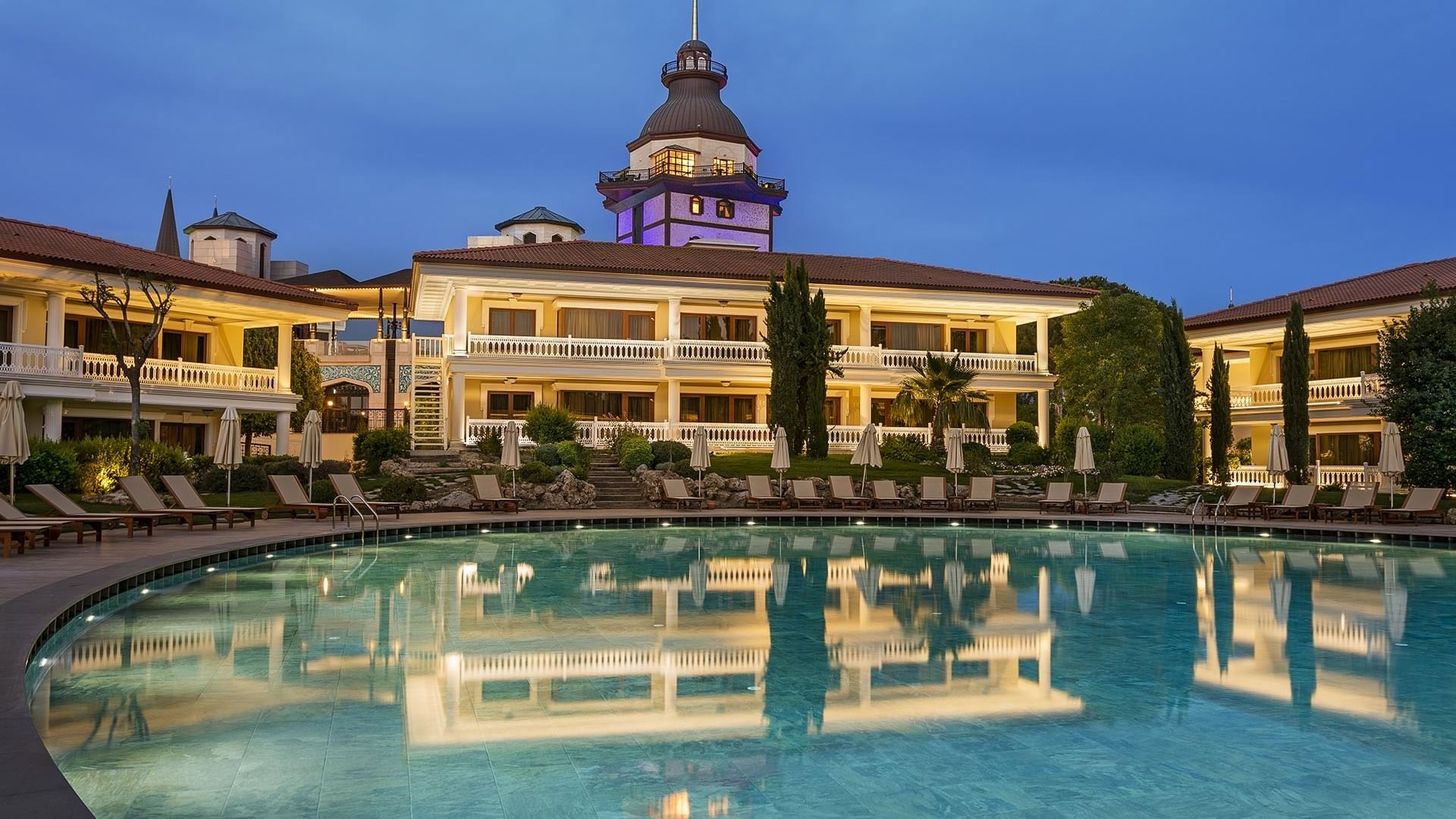 Luxuriöses Lake House mit Pool und Liegestühlen zwischen Bäumen unter dem nächtlichen Himmel.