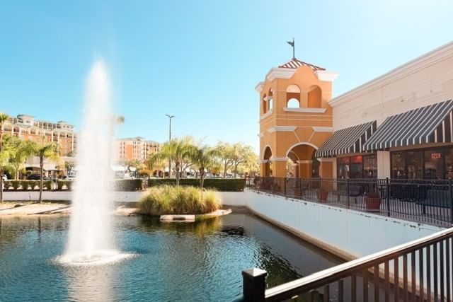 A large fountain and pond surrounded by parking lot and stucco storefronts. 