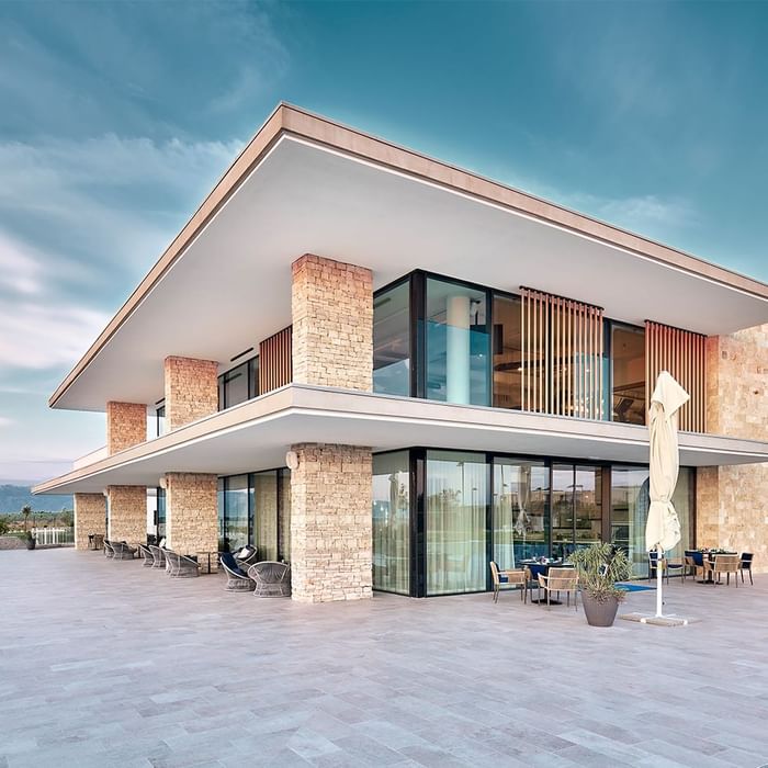 Moderne Villa mit Glasfassade, Terrasse und Möbeln unter blauem Himmel mit Wolken.