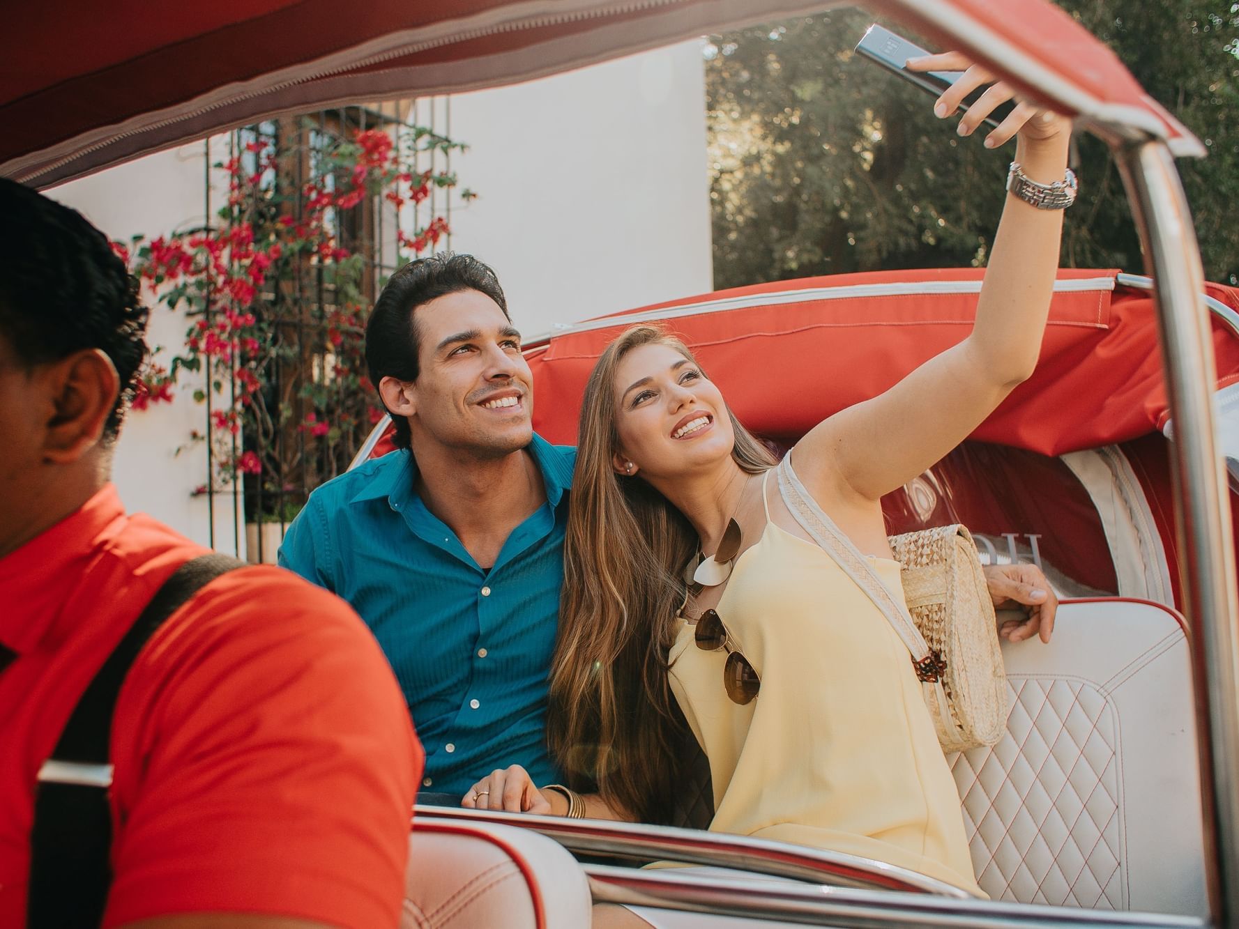 Couple taking a selfie while seated in a vintage car with vibrant flowers in the background at Billini Hotel Historic Luxury