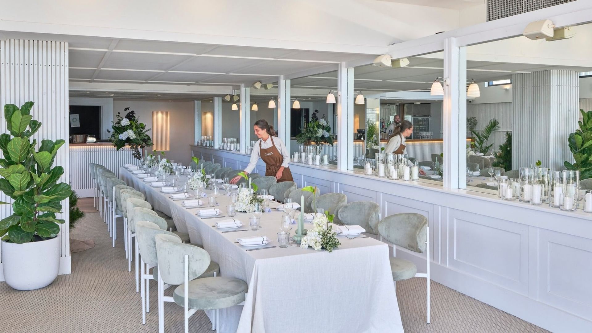 Staff member at Elysium Manly arranging a table surrounded by light grey chairs, near mirrors in the Cibaria\'s Terrazza