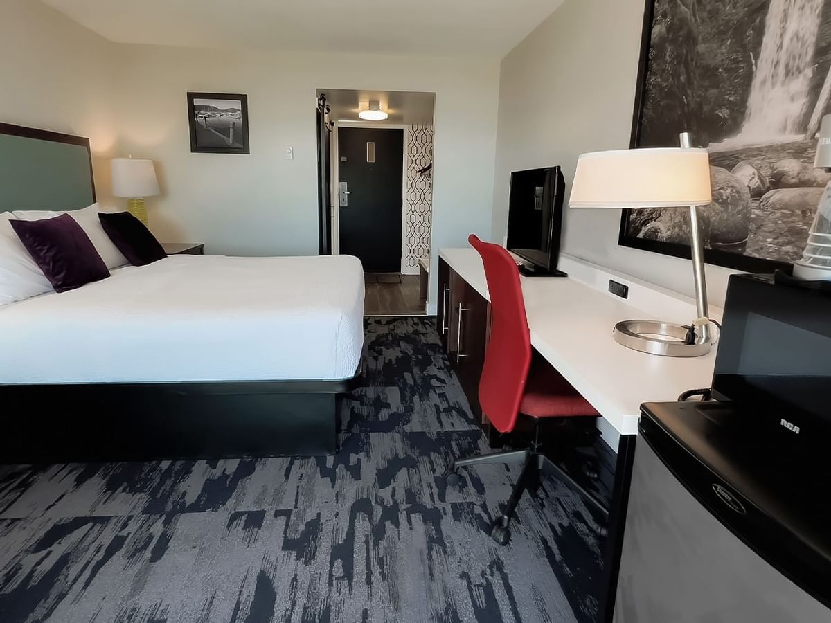 Guest room with king bed, lamps, red chair, desk, desk chair, microwave, fridge