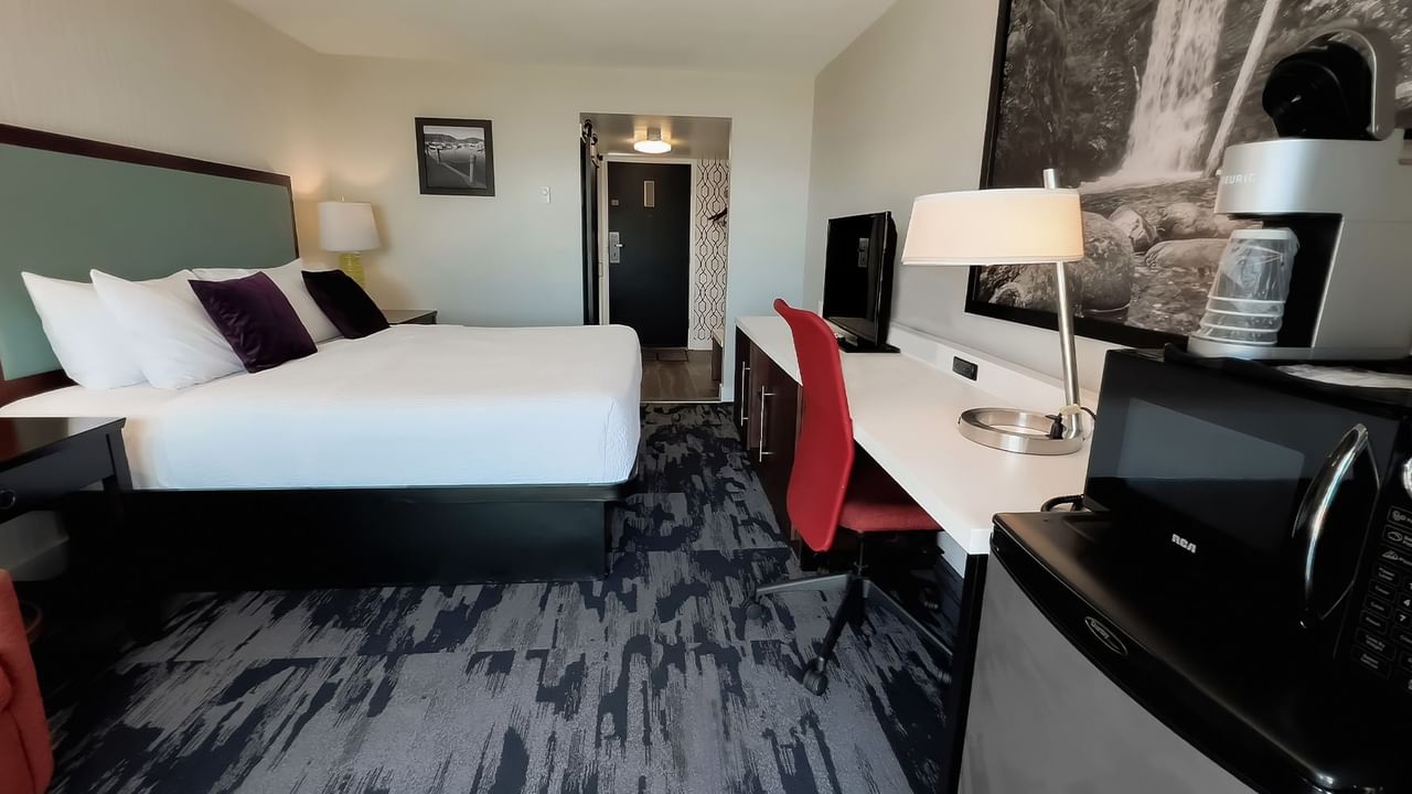 Guest room with king bed, lamps, red chair, desk, desk chair, microwave, fridge