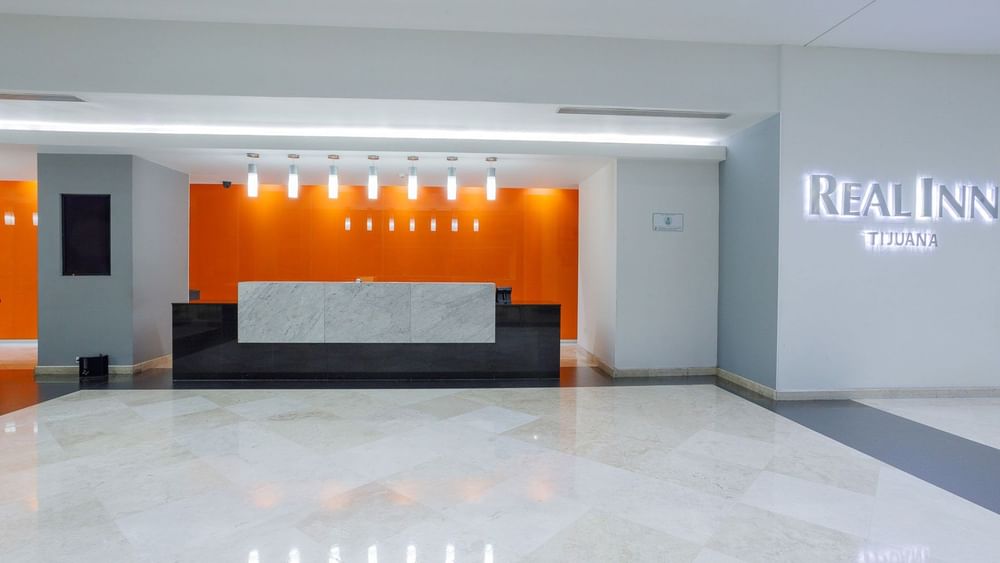 Reception desk set against an orange wall beneath pendant lights, & the brand sign displayed on a wall at Real Inn Tijuana