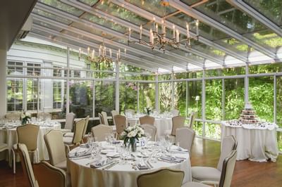 Round event tables with white tablecloths under a glass roof, chandeliers in glass room at Avon Old Farms Hotel