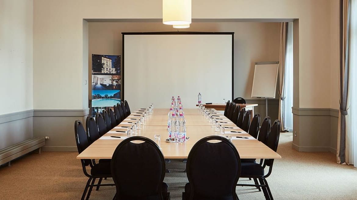 Salle de réunion avec tables, chaises, écran, tableaux blancs et fenêtres avec rideaux.