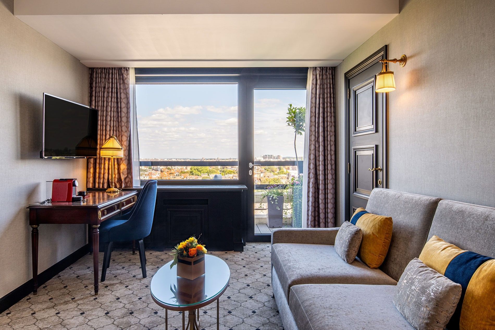 Grey sofa and work desk & wall-mounted TV with scenic city view from the terrace at Hotel Barsey by Warwick
