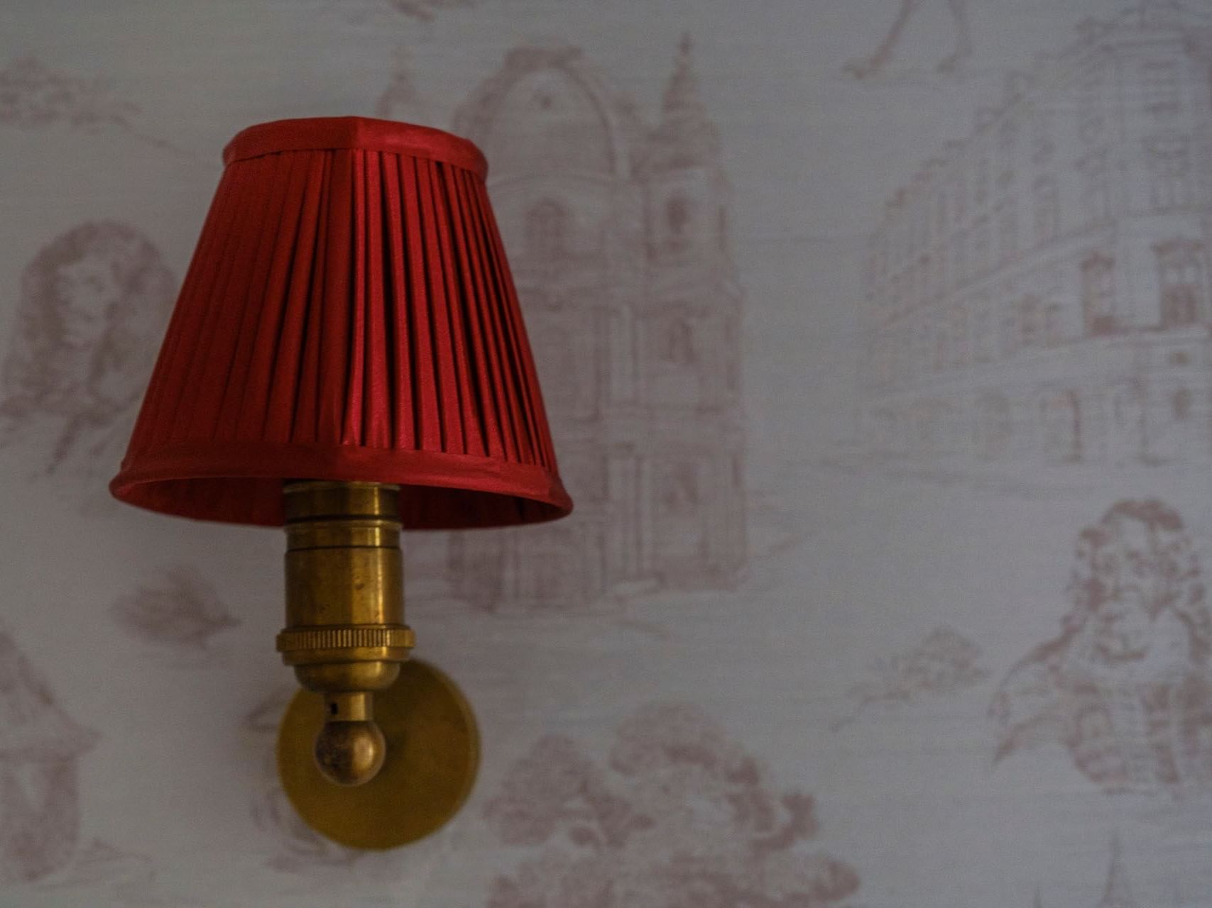 A gold wall lamp with a red pleated shade mounted on a white wall with a printed design.