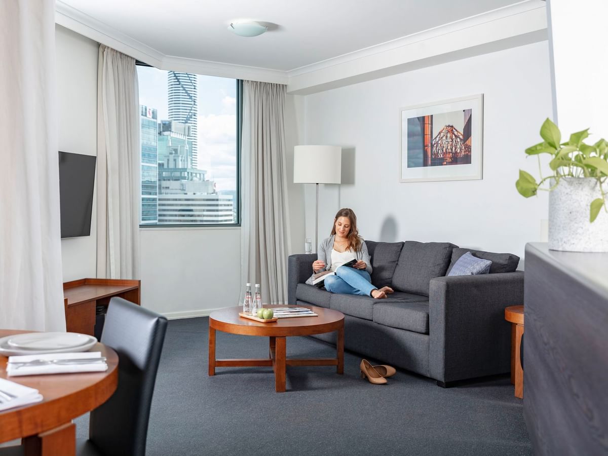 Woman reading a book on a dark sofa in Premium One Bedroom Apartments with a window view at The Sebel Brisbane