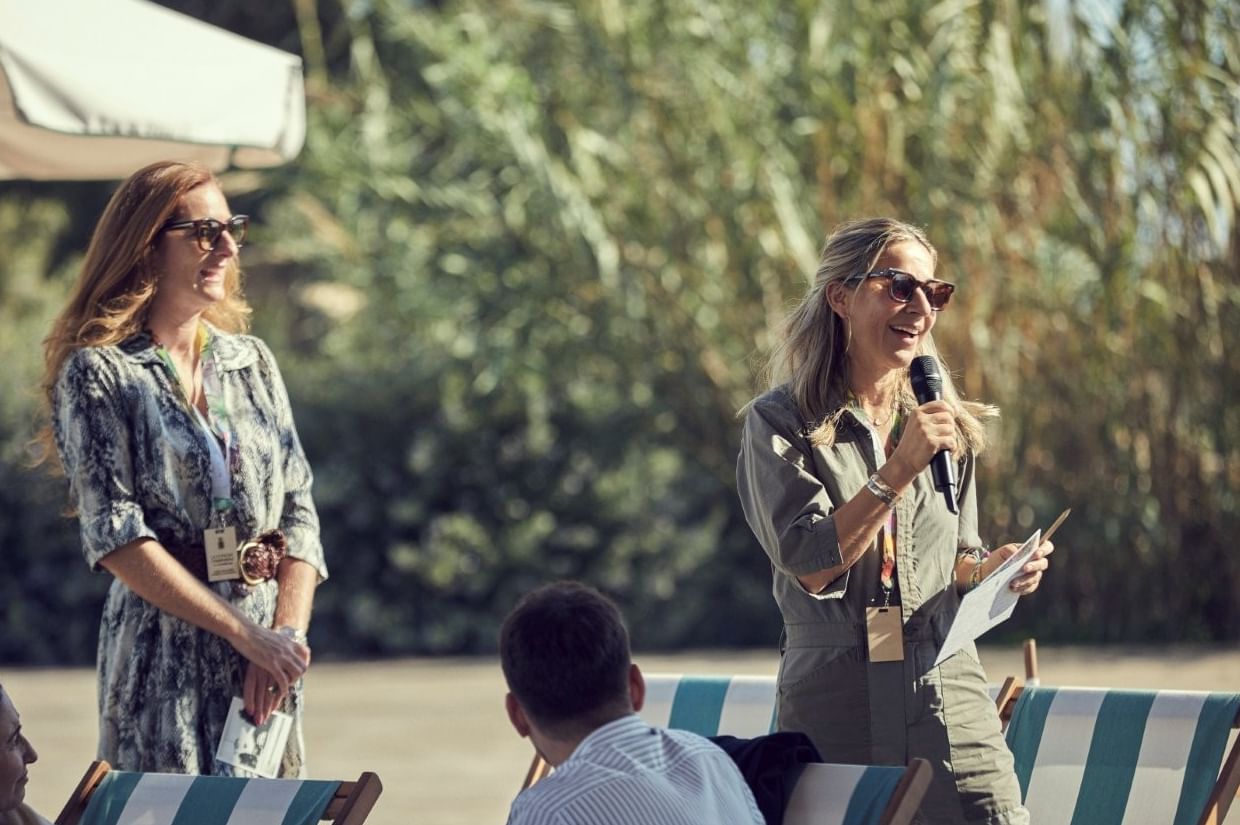 Woman in a jumpsuit and sunglasses speaking into a microphone outdoors at La Concha Conversations, Marbella Club