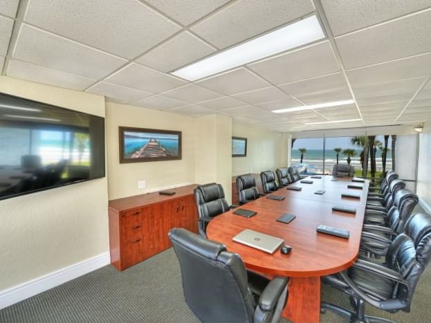 A large, oval-shaped wooden table with black leather chairs, a flat screen TV on the wall, and a beach view out the window.