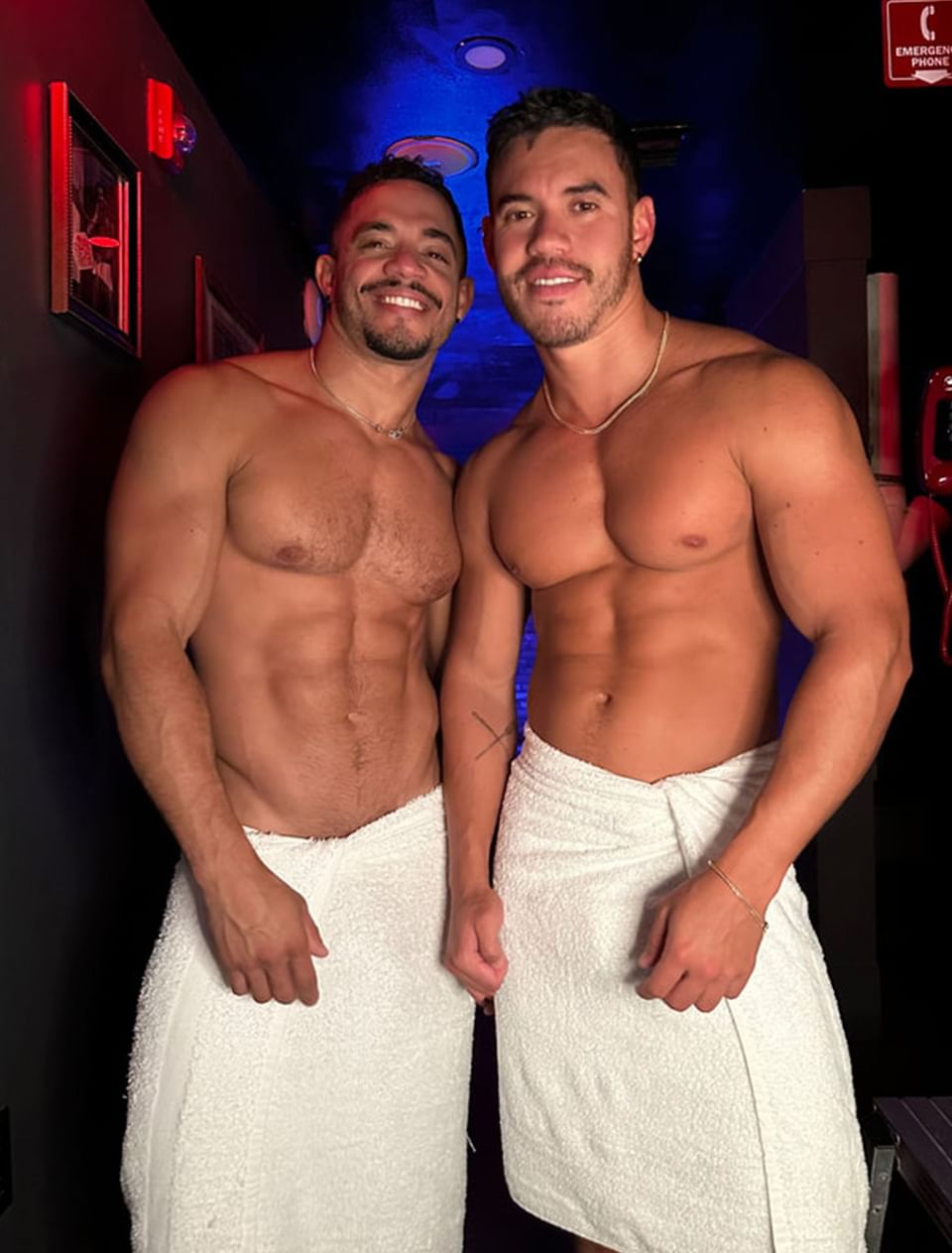 Two fit men pose shirtless in towels, showcasing toned physiques against a dimly lit, colorful background at The Gaythering