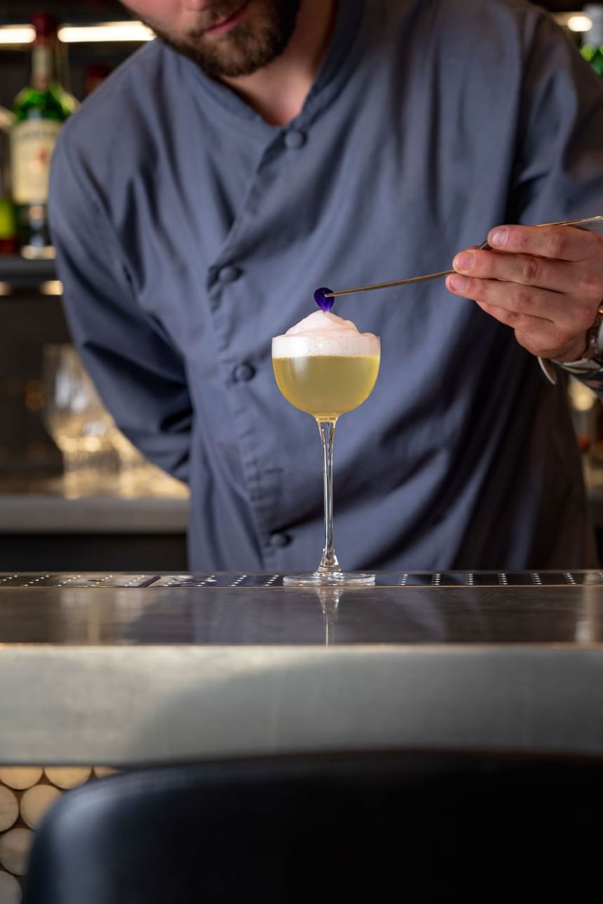 Bartender delicately adds a purple garnish to a frothy cocktail in a tall glass on a bar countertop at The Kitchen