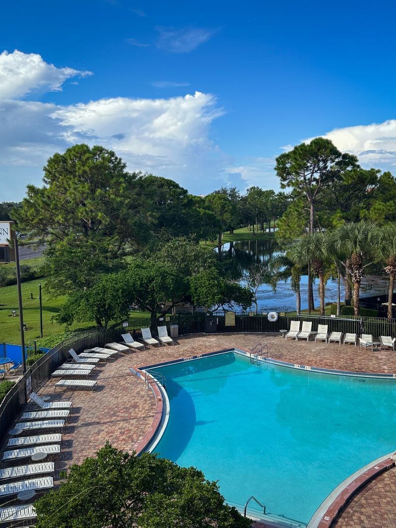 A view of the outdoor pool area at the Rosen Inn Lake Buena Vista, featuring a blue pool surrounded by white lounge chairs, lush green trees, and a bright blue sky with white clouds. 