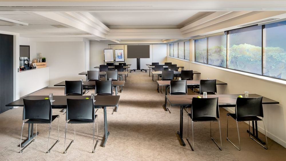 Classroom table setup facing the projector screen by the large windows in Acacia at Novotel Sydney International Airport