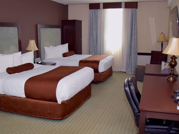 A hotel room with two beds, a desk, chair, lamp, TV, and window with curtains.