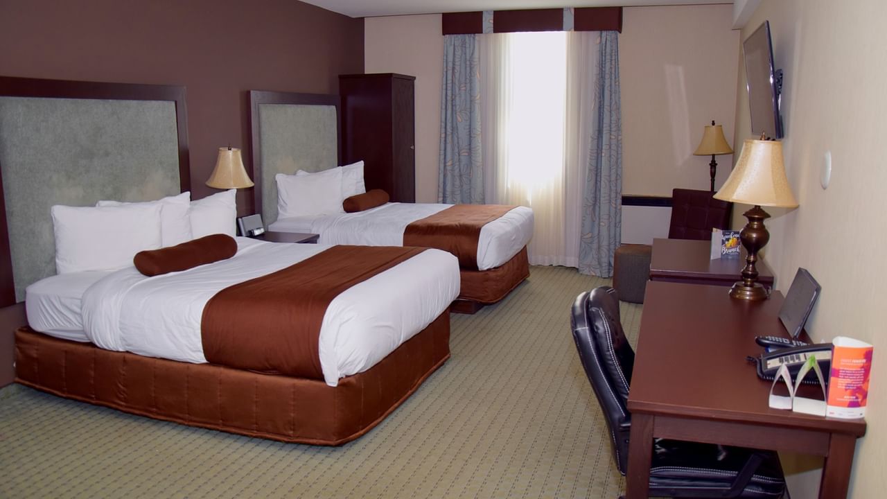 A hotel room with two beds, a desk, chair, lamp, TV, and window with curtains.