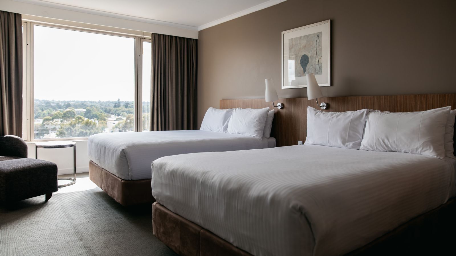 Two beds with white linens in a hotel room with large window and city view.