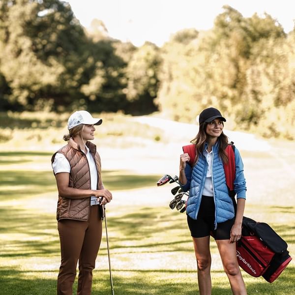 Due donne golfiste in abbigliamento sportivo, una con bastoni da golf e borsa, si parlano su un campo da golf.