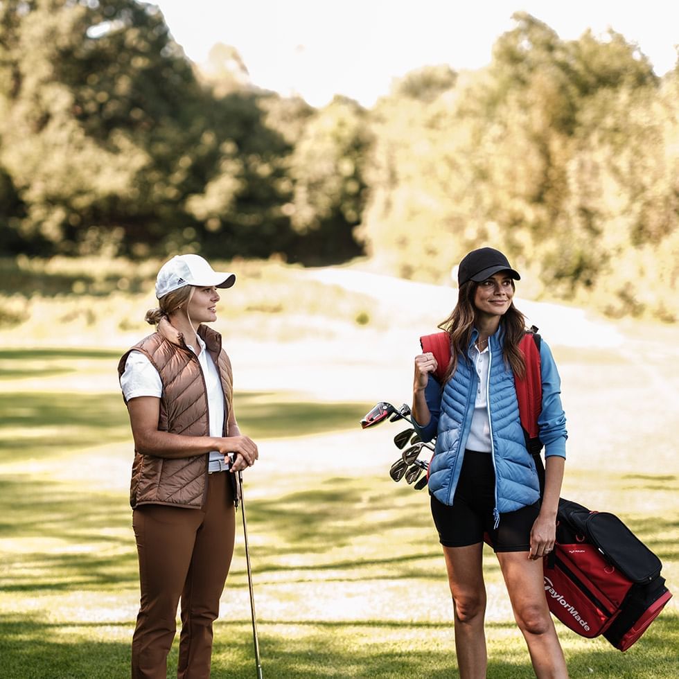Due donne con abbigliamento da golf e borse da golf in un campo erboso con alberi sullo sfondo.
