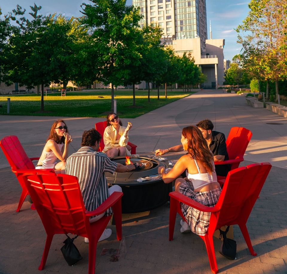 Group of young adults sitting in red chairs around a burning fire pit on a large paved courtyard at Hotel X Toronto