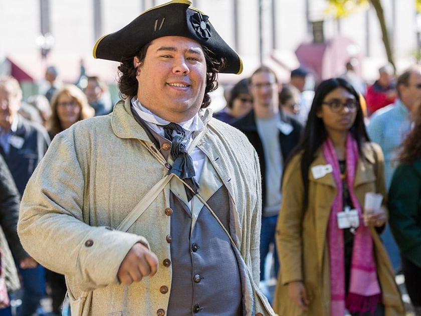 A man in colonial attire stands among a crowd of people, smiling and gesturing.