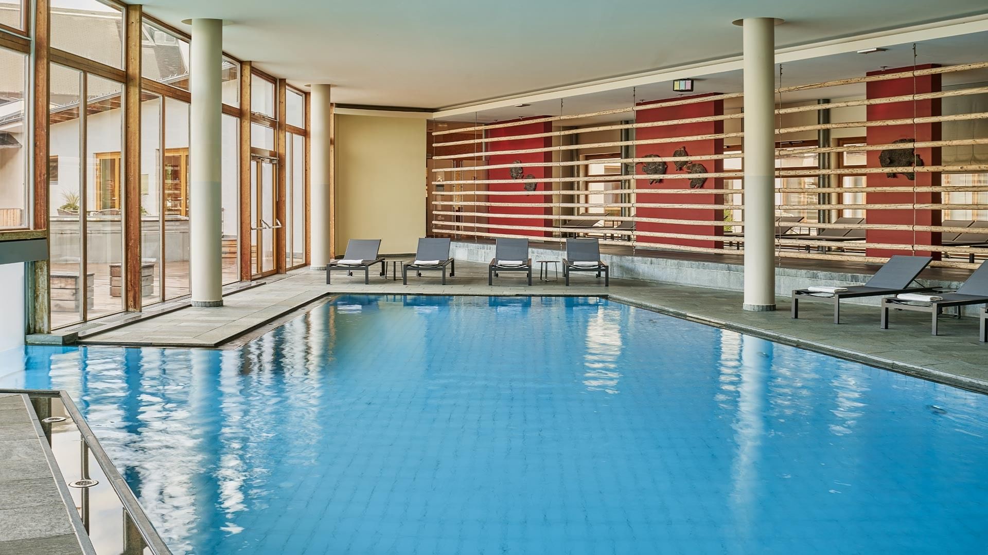 Empty indoor pool with lounge chairs, glass walls, and wooden partitions in a modern facility.