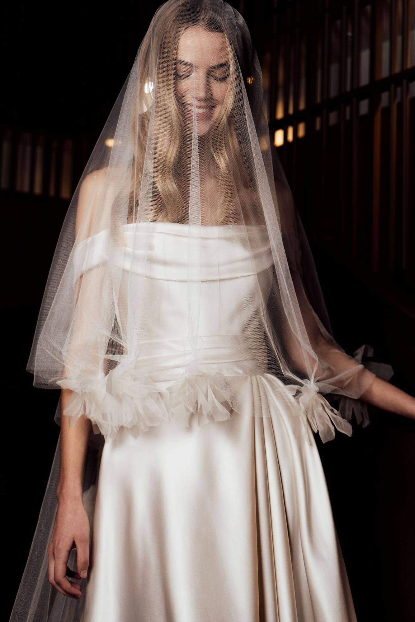 Bride smiling in a silk white wedding dress with a soft veil in dark background at The Londoner Hotel