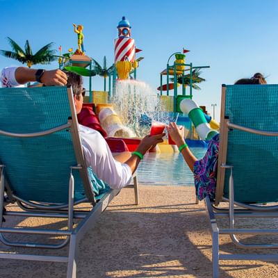 A couple relaxing in lounge chairs, toasting with drinks, with the splash pad and waterslides at Centennial Plaza Resort