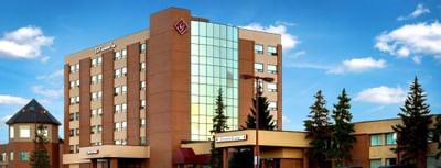 Exterior of The Glenmore Inn & Convention Centre, a tan high-rise with a glass atrium tower, pine trees under a blue sky