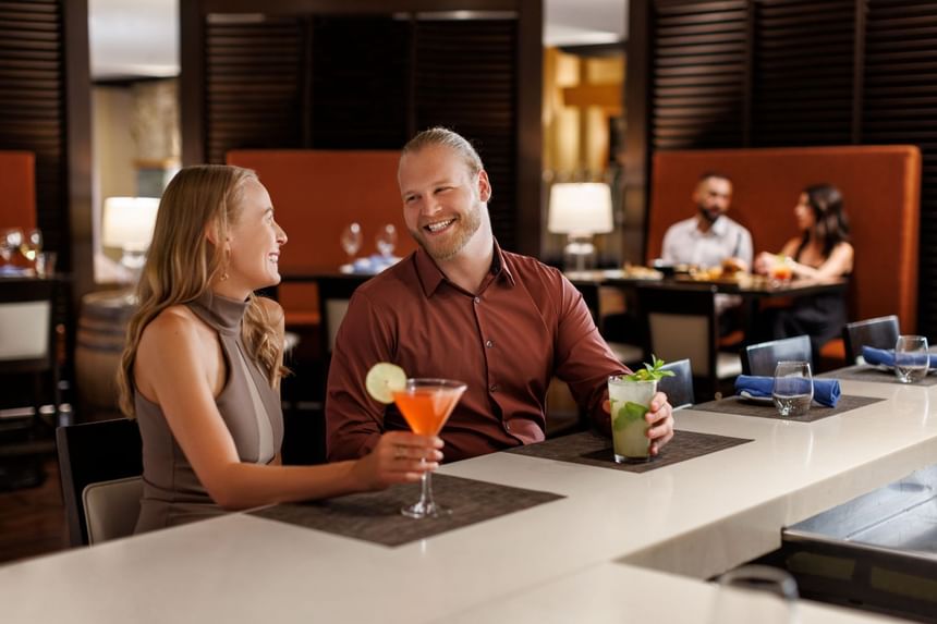 Two people sit at a bar, one with a cocktail, smiling, while others dine at tables behind them.
