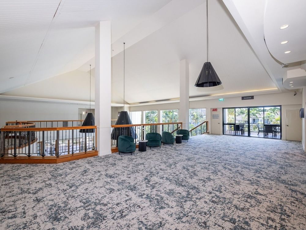 Empty lobby with green chairs, wooden railing, patterned carpet, and large glass doors leading to an outdoor area.