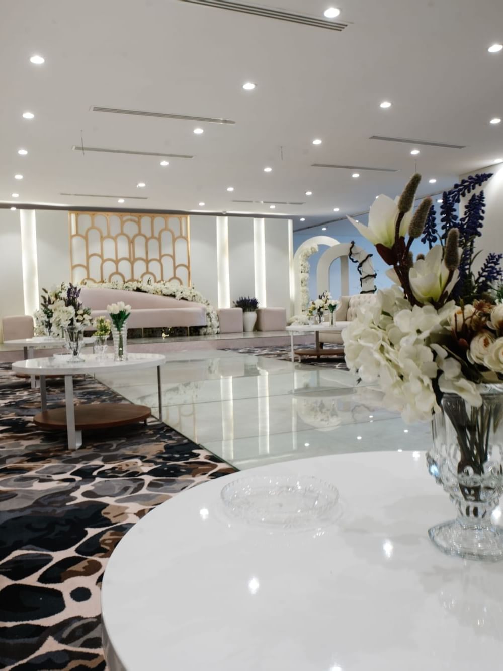 White floral vases by round tables in a bright hall, surrounding a plush white sofa at Cantonal Hotel by Warwick Riyadh